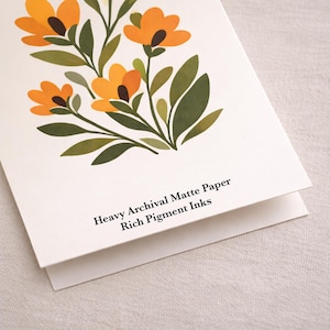 May include: A white greeting card featuring a botanical illustration of orange flowers with dark brown centers and green leaves. The text printed on the card reads: Heavy Archival Matte Paper, Rich Pigment Inks. The card is folded and matte.