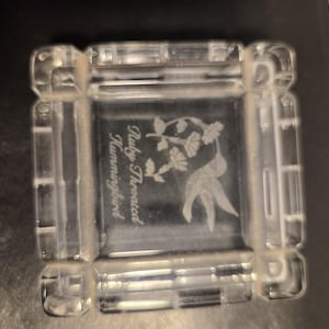 May include: A square clear glass paperweight featuring an etched design of a hummingbird hovering near a flower. The text Ruby Throated Hummingbird is engraved on the surface. The edges of the glass block have a faceted, geometric border design.