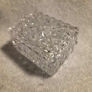 May include: A clear, rectangular glass trinket box featuring a faceted, diamond-cut pattern across the lid and sides. The box has a hinged lid with a geometric, star-like design on the top surface, reflecting light through its textured glass.
