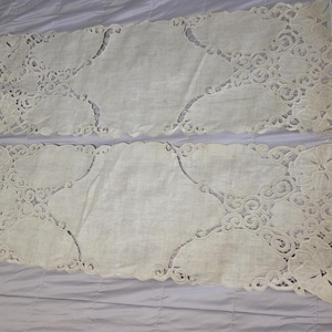 May include: Two off-white, rectangular table runners with intricate, openwork floral and scroll designs along the edges. The runners are laid out on a light purple surface, showcasing their delicate patterns and textures.