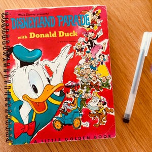 May include: A spiral-bound book titled Walt Disney presents Disneyland Parade with Donald Duck, featuring a large illustration of Donald Duck and various Disney characters on a red background, accompanied by a clear plastic ballpoint pen.