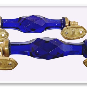 May include: Two cobalt blue glass door handles with brass accents. The handles are faceted and have a geometric design. The brass accents are in the shape of a rectangle with two holes.