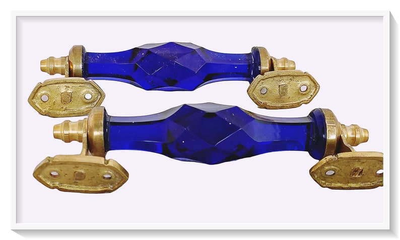 May include: Two cobalt blue glass door handles with brass accents. The handles are faceted and have a geometric design. The brass accents are in the shape of a rectangle with two holes.