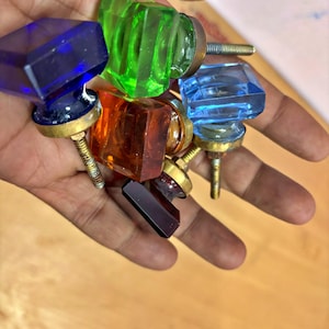 May include: A selection of colourful glass cabinet knobs in shades of blue, green, orange, and red. Each knob features a faceted, square design and a brass base with a threaded screw for installation. These knobs are a decorative hardware item.