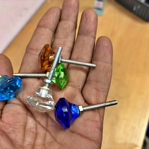 May include: A collection of colorful, faceted glass cabinet knobs in blue, clear, green, orange, and dark blue. Each knob is attached to a silver threaded rod. The knobs are held in a hand against a light wood surface.