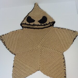 May include: A tan-colored, star-shaped hooded poncho with black stitched edges and a black face design on the hood. The hood features two black oval eyes and a stitched mouth. The poncho is made of a knitted material.