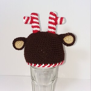 May include: A brown crocheted reindeer hat with red and white striped antlers and tan ears. The hat is placed on a clear glass. The bottom of the hat has a red and white striped trim. The hat is handmade.