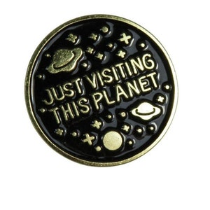 Pode incluir: Um broche de esmalte preto com letras douradas que diz "Just Visiting This Planet". O broche tem um contorno dourado e um desenho de planetas e estrelas.