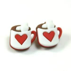 May include: A pair of white ceramic earrings shaped like mugs with red hearts and brown rims. Each mug has a white swirl on top, resembling whipped cream.