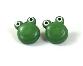 May include: Two green frog-shaped buttons with white eyes and black pupils. The frogs are smiling.