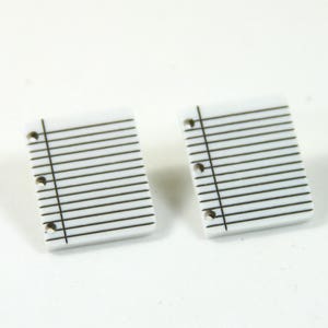 May include: A pair of white, paper-like earrings with a lined pattern and holes for a binder. The earrings are small and would be a fun and quirky accessory.