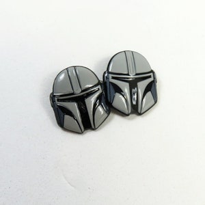 Puede incluir: Dos pines de esmalte de casco mandaloriano en plata y negro. Los pines están detallados con una visera negra y un casco plateado.