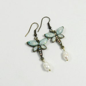 Puede incluir: Un par de pendientes colgantes con un diseño de libélula con alas azules iridiscentes y una gota de perla. La libélula está adornada con pedrería.