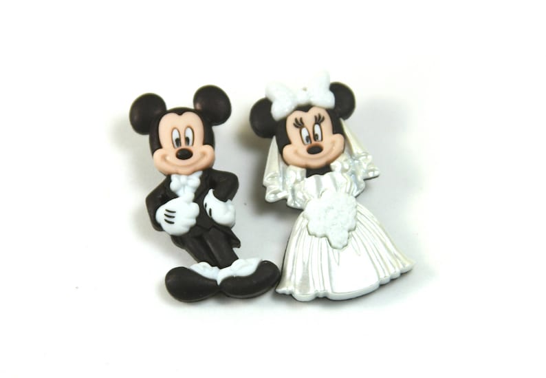 May include: A pair of plastic figurines of Mickey Mouse and Minnie Mouse dressed in wedding attire. Mickey is wearing a black tuxedo with a white bow tie and Minnie is wearing a white wedding dress with a veil.