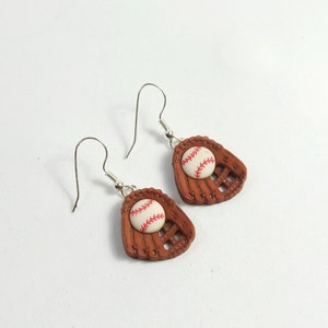 Könnte beinhalten: Ein Paar silberne Ohrhänger mit braunen Baseballhandschuh-Anhängern und weißen Baseballs mit roten Nähten.