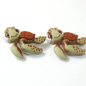May include: Two green and brown turtle figurines with white eyes and smiling mouths. The turtles are lying on their backs with their flippers outstretched.