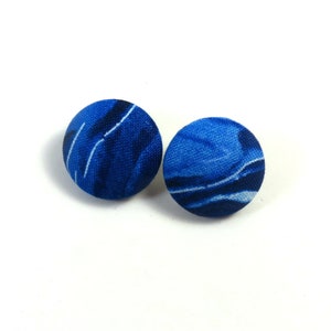 May include: Two round fabric-covered buttons with a blue and white abstract pattern. The buttons are a deep shade of blue with white streaks, creating a marbled effect. They are set against a plain white background.