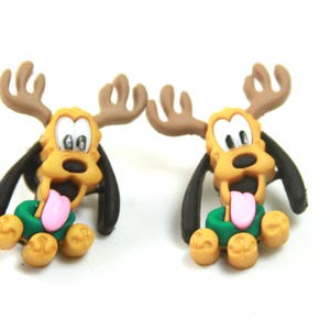 May include: A pair of brown and tan reindeer dog earrings with green and gold accents. The dog is wearing a green scarf and has a pink tongue sticking out.
