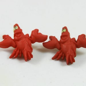 Puede incluir: Dos figuritas de plástico rojo de Sebastián el cangrejo de La Sirenita de Disney. Las figuritas son de estilo de dibujos animados con ojos grandes y una amplia sonrisa.