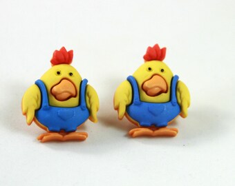 May include: Two yellow chick-shaped buttons with orange beaks and feet, and red crests. They wear blue dungarees. The buttons are set against a white background, creating a cheerful and playful aesthetic.