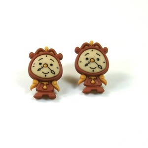 May include: A pair of brown and gold-colored earrings featuring a cartoon character of a clock with a face. The clock character has a brown body, a gold face, and a gold crown on its head.