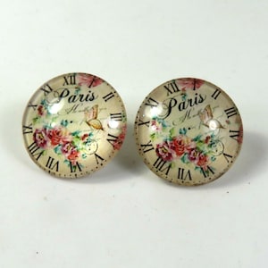 May include: A pair of round glass earrings with a vintage Parisian clock face design. The clock face features Roman numerals, a butterfly, and the words "Paris" and "Amour". The background is a floral pattern with pink and white roses.