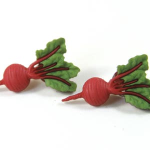 May include: A pair of red and green beet earrings. The beets are realistic and have green leaves.