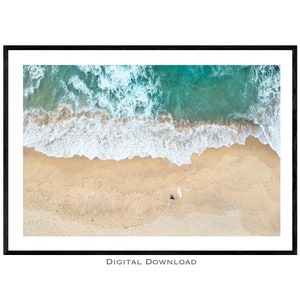 May include: An aerial view of a sandy beach with turquoise water and white waves crashing on the shore. A lone person is walking on the beach with a surfboard.