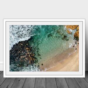 May include: An aerial view of a rocky coastline with turquoise water and a sandy beach. The waves are crashing against the rocks, creating a foamy white border.