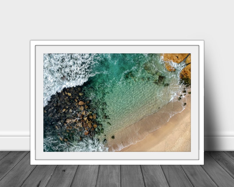 May include: An aerial view of a rocky coastline with turquoise water and a sandy beach. The waves are crashing against the rocks, creating a foamy white border.