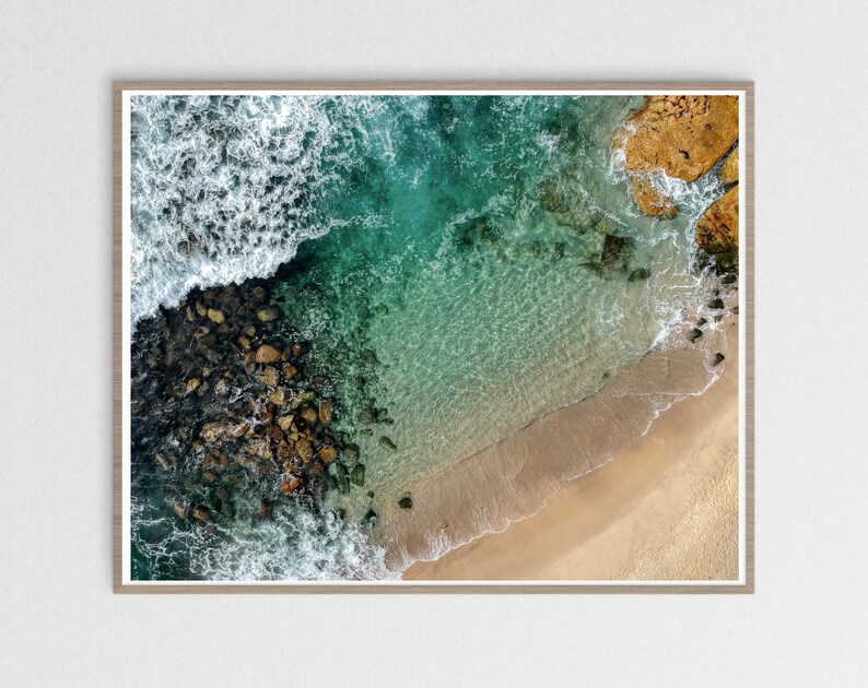 May include: An aerial view of a rocky coastline with turquoise water crashing against the shore. The water is clear and shallow, revealing the sandy bottom. The rocks are a mix of brown and gray, and the sand is a light beige.