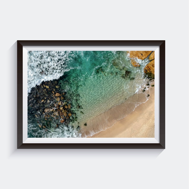 May include: An aerial view of a rocky coastline with turquoise water and a sandy beach. The waves are crashing against the rocks, creating a foamy white border.