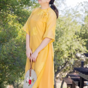 May include: A woman in a yellow dress with a floral pattern holds a small, round, embroidered bag with a brown strap. The bag has pink and red flowers embroidered on it.