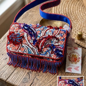 May include: A rectangular handbag featuring intricate beadwork and sequins in red, blue, and orange floral and paisley patterns. The bag includes a blue strap, a beaded fringe along the bottom edge, and a white tag labeled Two Coda Sisters.