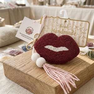 May include: A beaded keychain shaped like lips in deep red with a white center, attached to a gold-tone metal clasp. It features two white spherical beads and a long, light pink beaded tassel. The item is displayed on a branded card with the text Bead-Handmade.