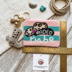 May include: A rectangular beaded coin purse with pink and teal stripes, featuring a leopard print cowboy hat and the text "RODEO babe". It includes a gold keychain with a matching leopard print boot charm, measured by a 7-inch ruler.