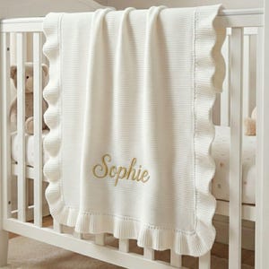 May include: A cream-coloured baby blanket with a ruffled edge and the name "Sophie" embroidered in gold thread. The blanket is draped over a white cot, with a teddy bear visible in the background. The blanket is made of a soft, knitted material.