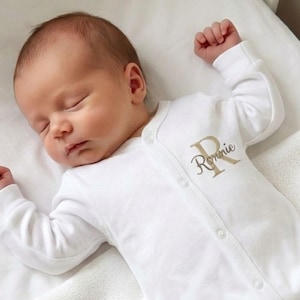 May include: A sleeping baby in a white onesie with long sleeves. The onesie features a gold embroidered initial "R" and the name "Ronnie". The baby is resting on a white surface.