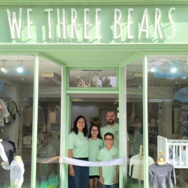 May include: A family stands in front of a store with the text "WE THREE BEARS" above the entrance. The store is painted a light green color. The family is wearing matching light green shirts. A white ribbon is stretched across the doorway.