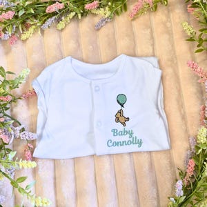 K&ouml;nnte beinhalten: Wei&szlig;er Baby-Body mit Knopfverschluss. Er hat ein gesticktes Design eines braunen B&auml;ren, der einen tealfarbenen Ballon h&auml;lt, mit dem Text "Baby Connolly" in tealfarbener Schrift. Der Body ist auf einer rosa-wei&szlig; gestreiften Oberfl&auml;che gefaltet, umgeben von Gr&uuml;n.