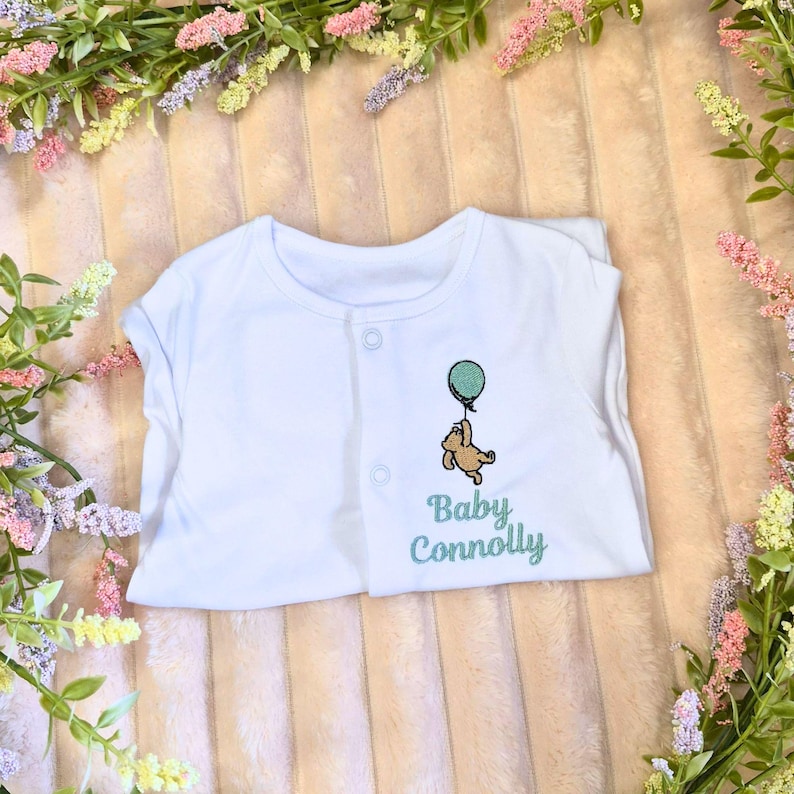 K&ouml;nnte beinhalten: Wei&szlig;er Baby-Body mit Knopfverschluss. Er hat ein gesticktes Design eines braunen B&auml;ren, der einen tealfarbenen Ballon h&auml;lt, mit dem Text "Baby Connolly" in tealfarbener Schrift. Der Body ist auf einer rosa-wei&szlig; gestreiften Oberfl&auml;che gefaltet, umgeben von Gr&uuml;n.