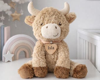 May include: A plush, light brown Highland cow stuffed animal with a heart-shaped wooden tag that reads "Isla". The toy has a cream-colored face, horns, and hooves. It is sitting on a white surface.