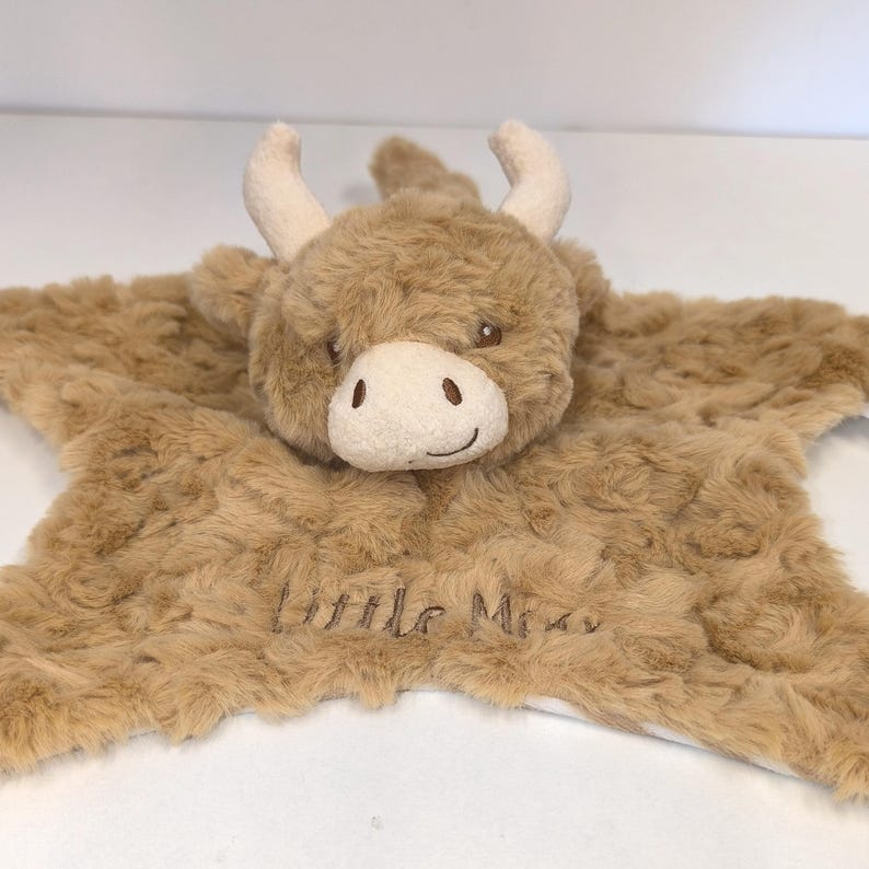 May include: A plush, brown Highland cow baby lovey. The cow has a white muzzle, small horns, and embroidered eyes. The lovey has a star-shaped body with the words "Little Miss" embroidered on it.