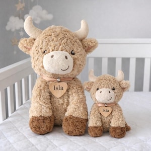 May include: Two soft Highland cow plush toys, one large and one small, with tan fur and brown hooves. Each cow has a heart-shaped wooden tag that reads "Isla" and a pink collar. The background features a white cot and a grey wall.