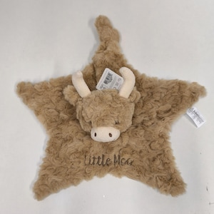 May include: A plush, star-shaped baby comforter in shades of brown, featuring a Highland cow face with cream-colored horns and a soft muzzle. The text "Little Moo" is embroidered on the comforter. Includes a product tag.