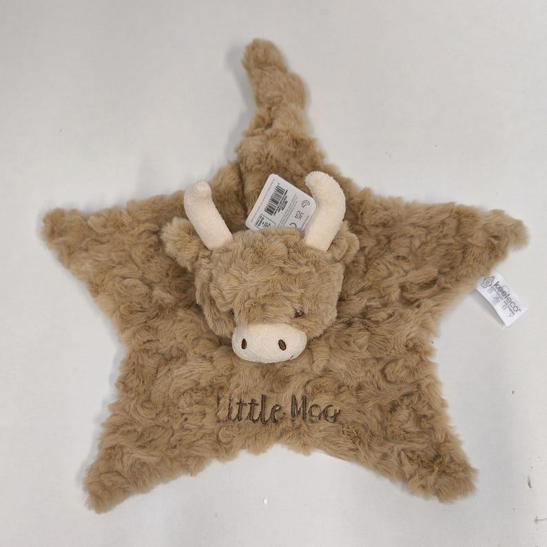 May include: A plush, star-shaped baby comforter in shades of brown, featuring a Highland cow face with cream-colored horns and a soft muzzle. The text "Little Moo" is embroidered on the comforter. Includes a product tag.