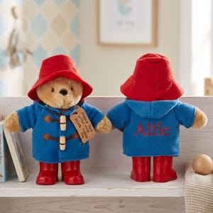 May include: Two Paddington Bear plush toys. Each bear is wearing a red hat, blue coat, and red wellington boots. One has a tag saying "Please look after this BEAR. THANK YOU." The other has "Alfie" embroidered on the back of the coat.
