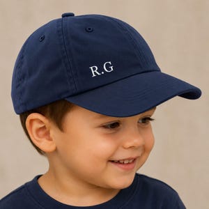 May include: A navy blue baseball cap featuring the white initials "R.G". The cap has a curved peak and a button on the crown. The cap is made of a soft fabric.