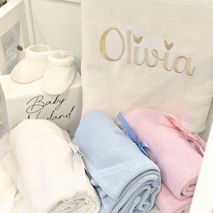 May include: A collection of baby items, including white knitted booties, a white box with "Baby Land" text, and rolled blankets in white, blue, and pink. A cream-colored blanket is embroidered with the name "Olivia" in gold.