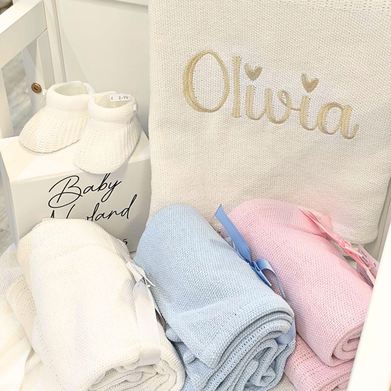 May include: A collection of baby items, including white knitted booties, a white box with "Baby Land" text, and rolled blankets in white, blue, and pink. A cream-colored blanket is embroidered with the name "Olivia" in gold.